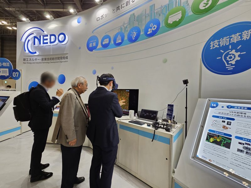 A visitor wearing a VR headset tries the VR co-creation environment at NEDO's booth while two others watch